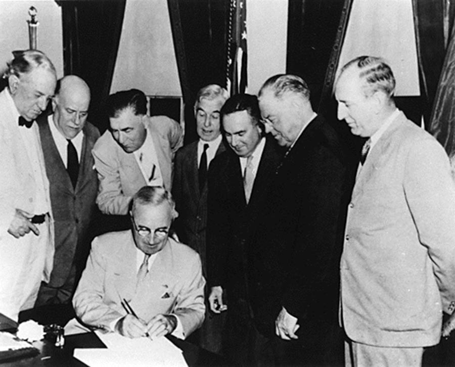 Black and white photo of President Truman surrounded by several men as he signs the Atomic Energy Act of 1946.