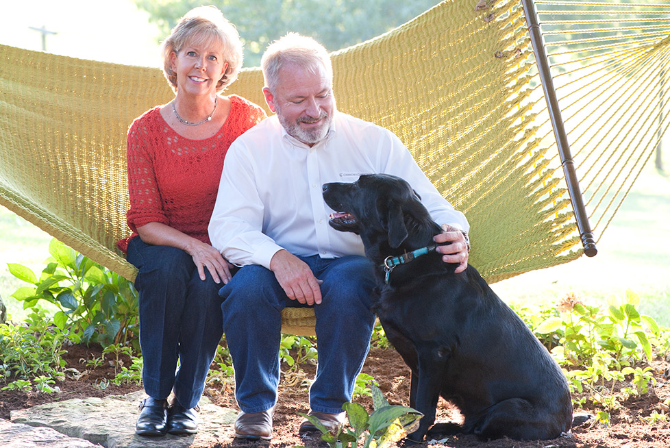 Angie and Harold Cannon sit on a hammock with their dog in front.