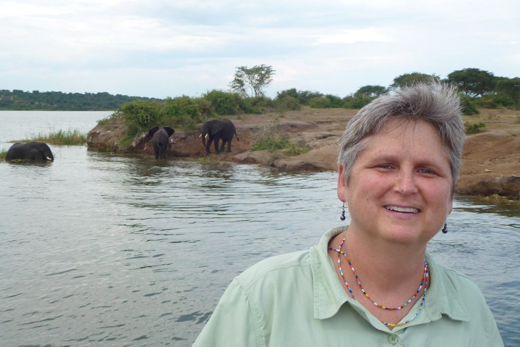 Laura Lackey poses with elephants on the other side of a body of water.